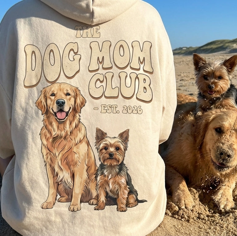 Person wearing a hoodie with 'Dog Mom Club' design on a beach with dogs.