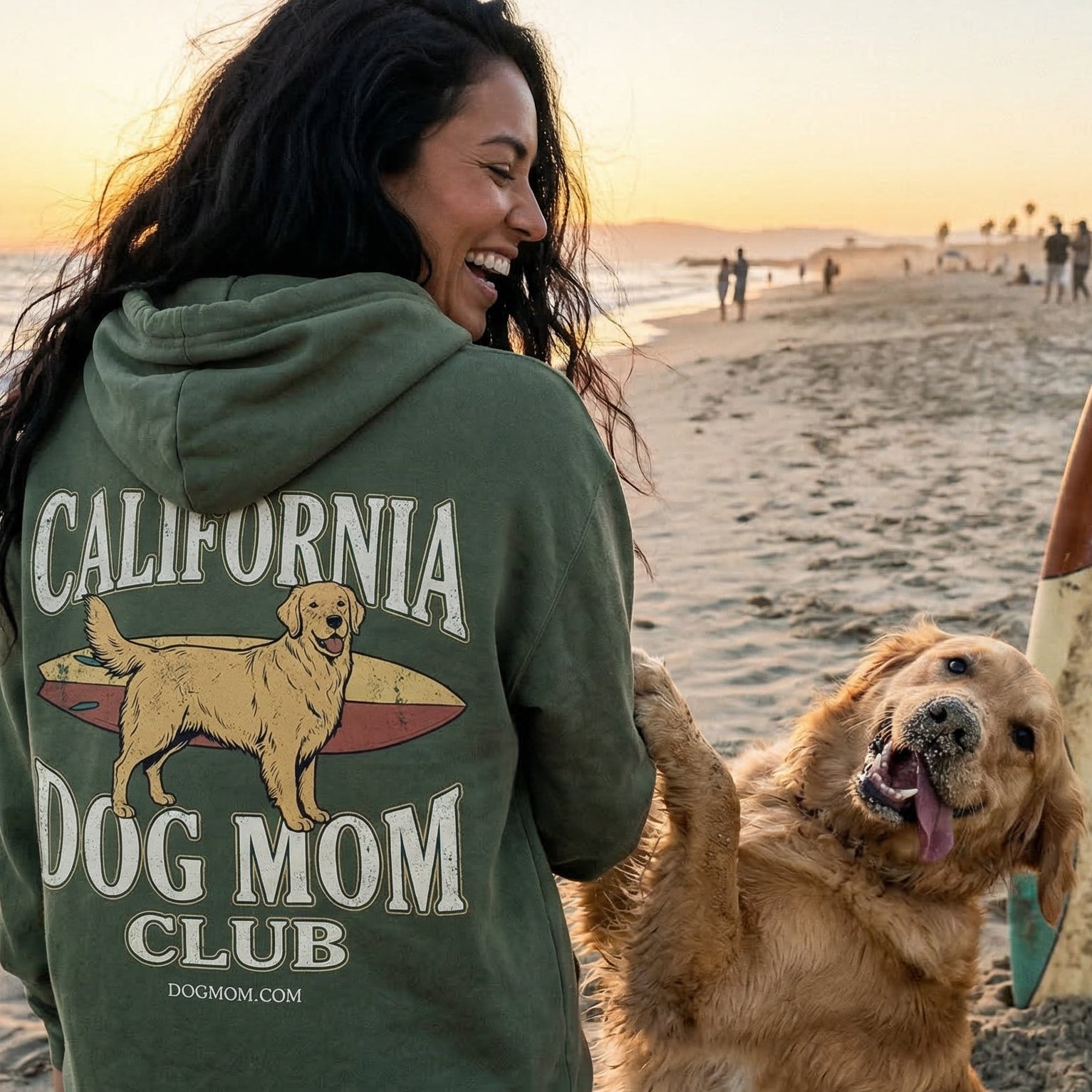 Woman wearing a 'California Dog Mom Club' hoodie on a beach with a dog and surfboard.