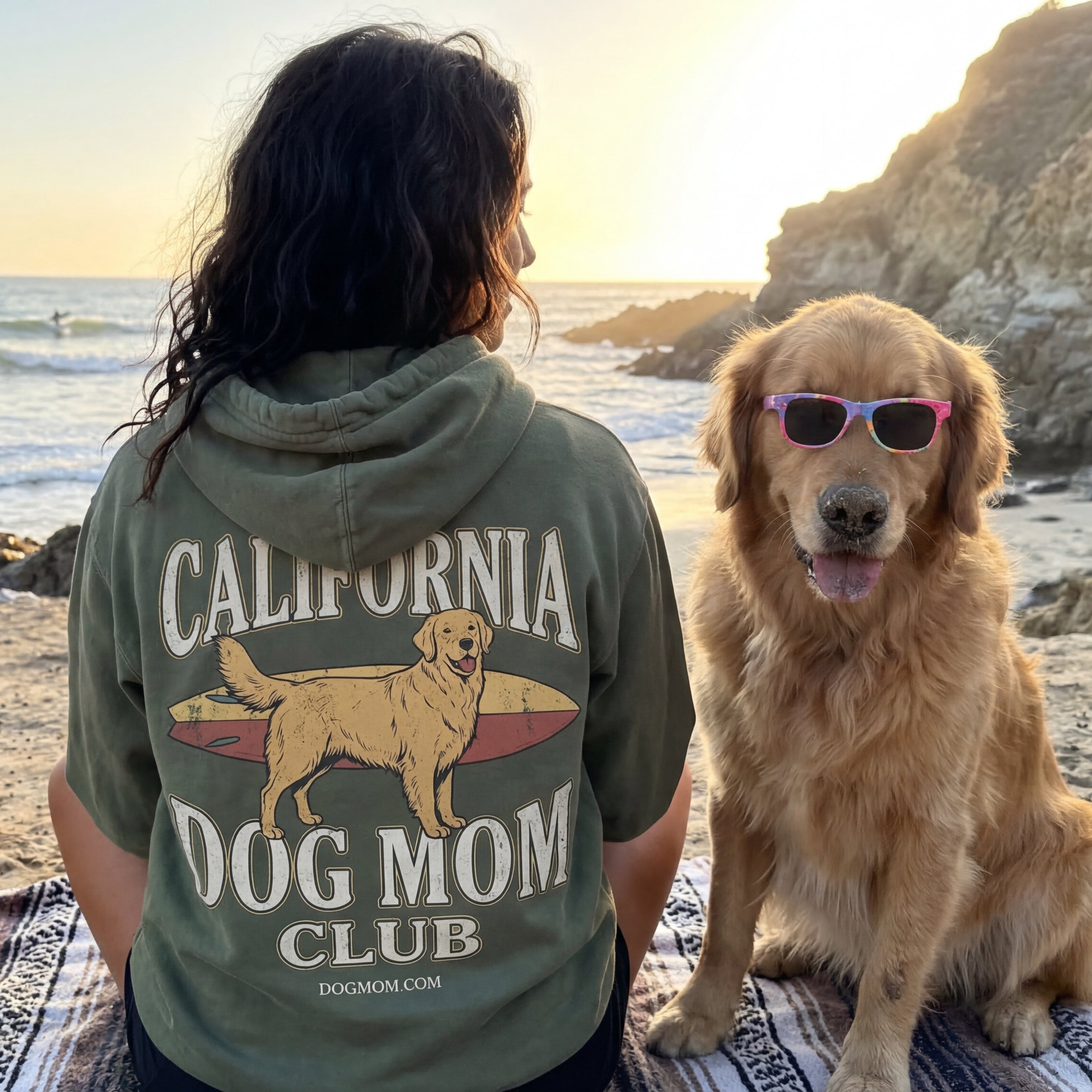 Person wearing a 'California Dog Mom Club' hoodie with a dog on a beach.