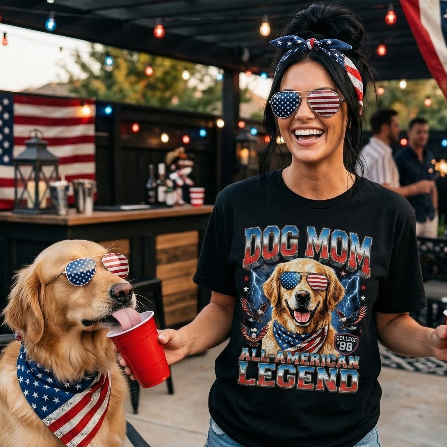 Woman wearing a 'Dog Mom All American Legend' shirt with a dog, both wearing sunglasses, at a festive outdoor event.