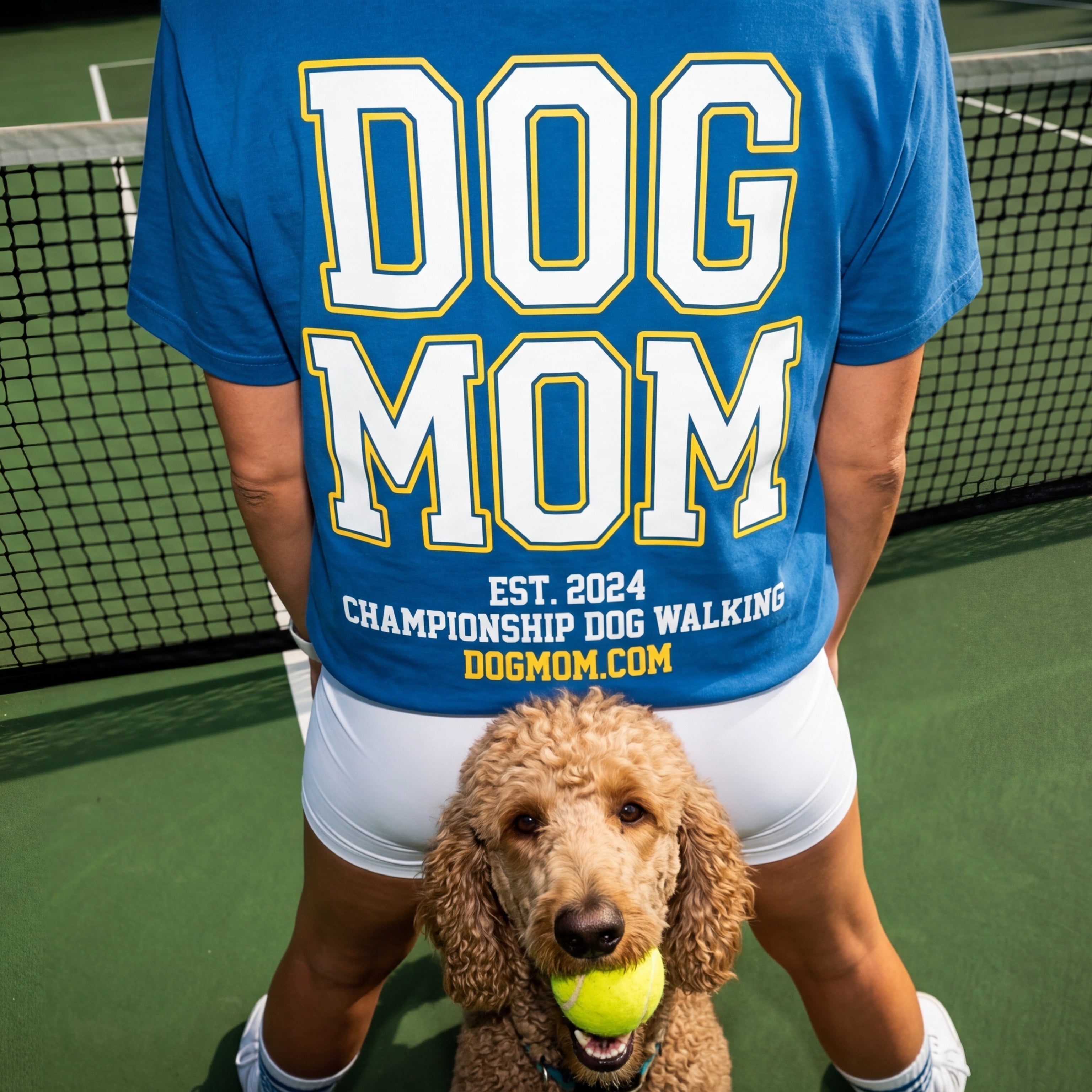 Person wearing a 'DOG MOM' shirt on a tennis court with a dog holding a tennis ball.