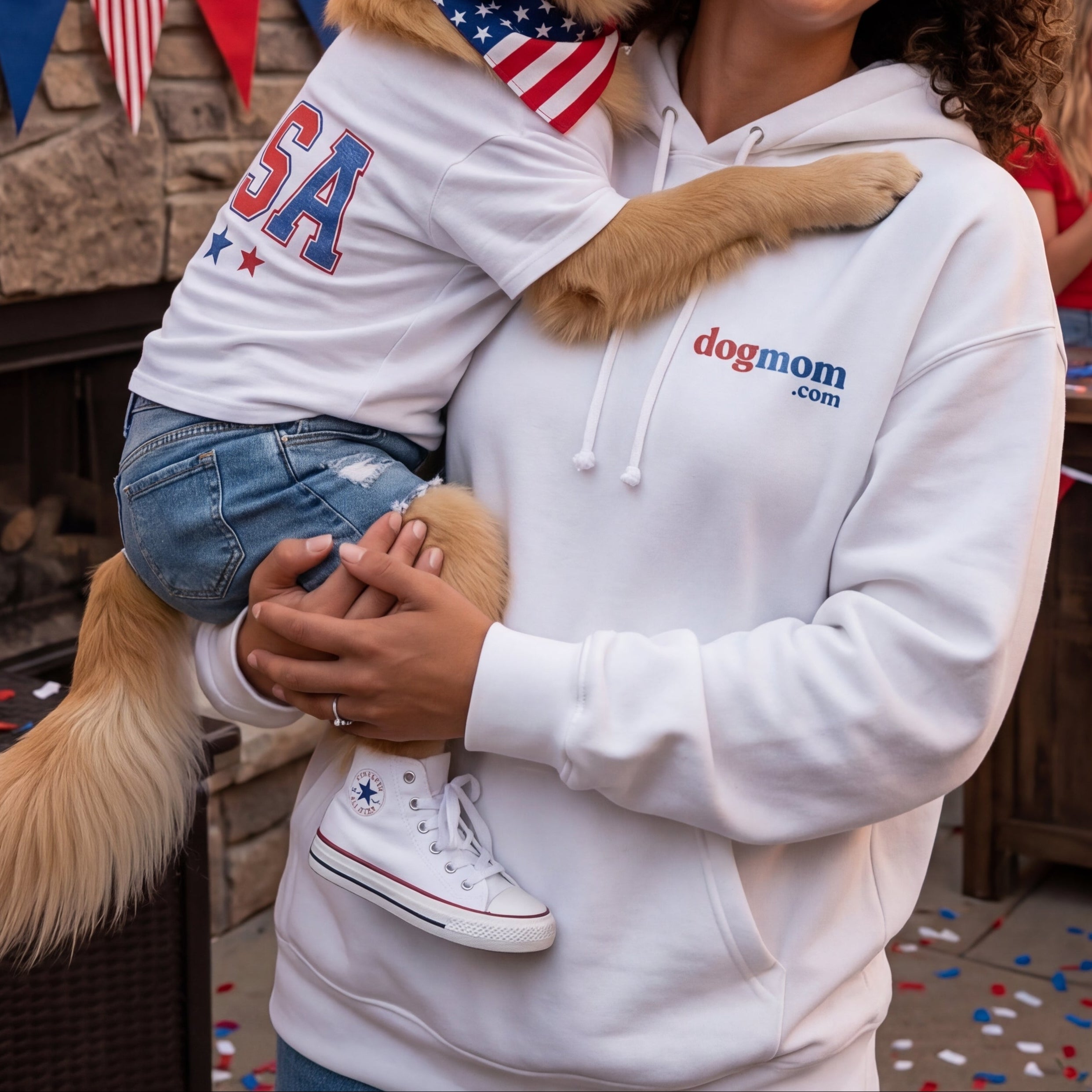 Woman holding a dog wearing a 'USA' hat with fireworks in the background, surrounded by people and decorations.